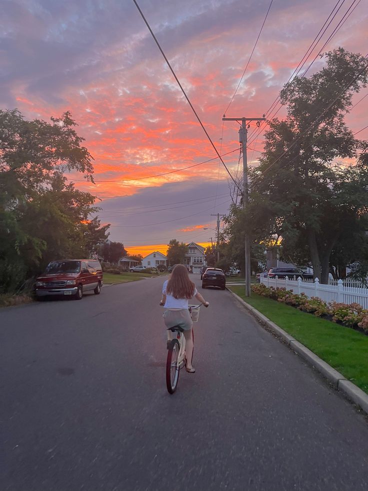 a woman is riding her bike down the street at sunset with cars in the background