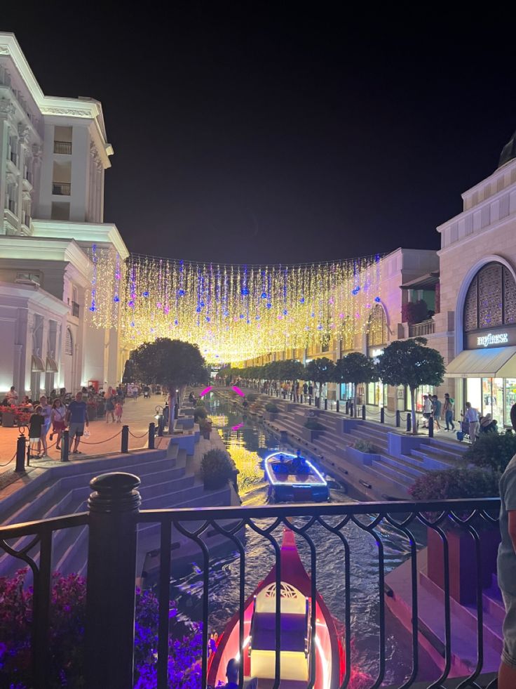 people are walking on the street at night with lights hanging from buildings in the background