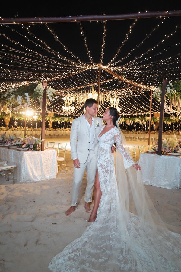 a bride and groom pose for a photo on the beach at night with lights in the background