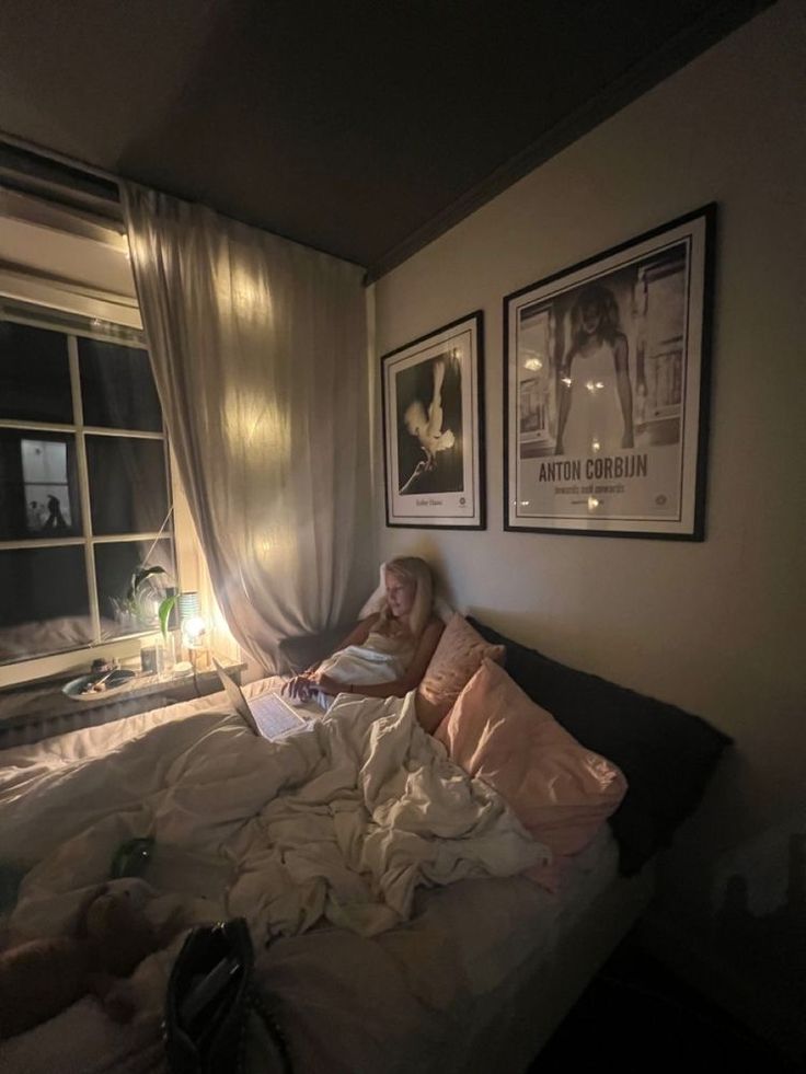 a woman laying in bed reading a book next to a window with pictures on the wall