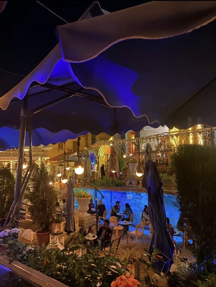 people sitting at tables under umbrellas in an outdoor area with lights on the buildings