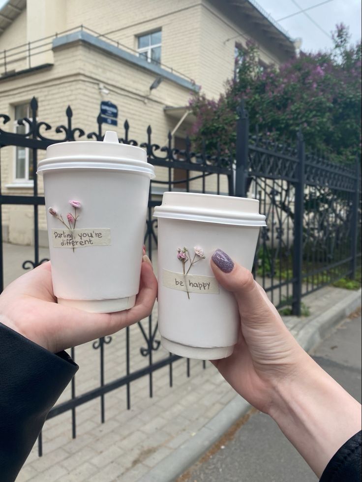 two people holding up coffee cups in front of a fence