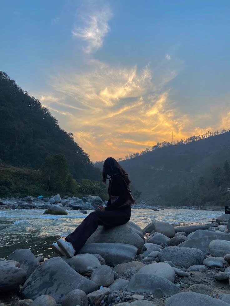 a woman sitting on top of a rock next to a river under a cloudy sky