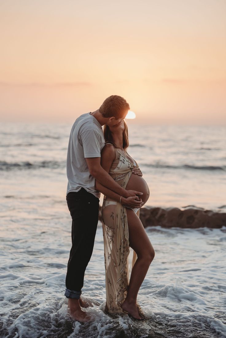 a pregnant couple kissing in the ocean at sunset with their belly wrapped around each other