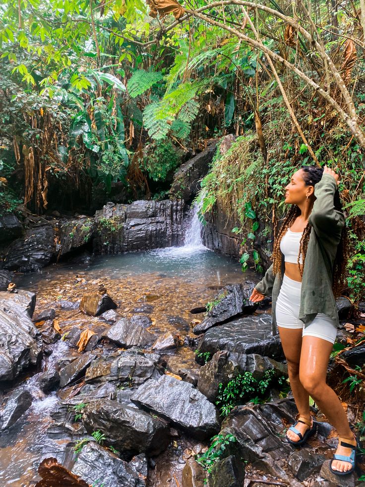 Black girl standing in rainforest with oversized green button down and cream activewear set. Waterfall in the background. She has passion twists. Nature Trip Outfit Summer, Puerto Rico Outfits Aesthetic, Vacation Hiking Outfits, Rainforest Outfits Women, Rainforest Vacation Outfits, Puerto Rico Nightlife Outfit, Tropical Hike Outfit, La Fortuna Costa Rica Outfits, El Salvador Aesthetic Outfit