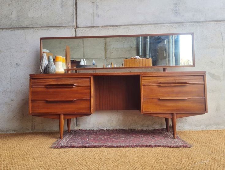 a wooden desk with two drawers and a large mirror on top of it, in front of a concrete wall