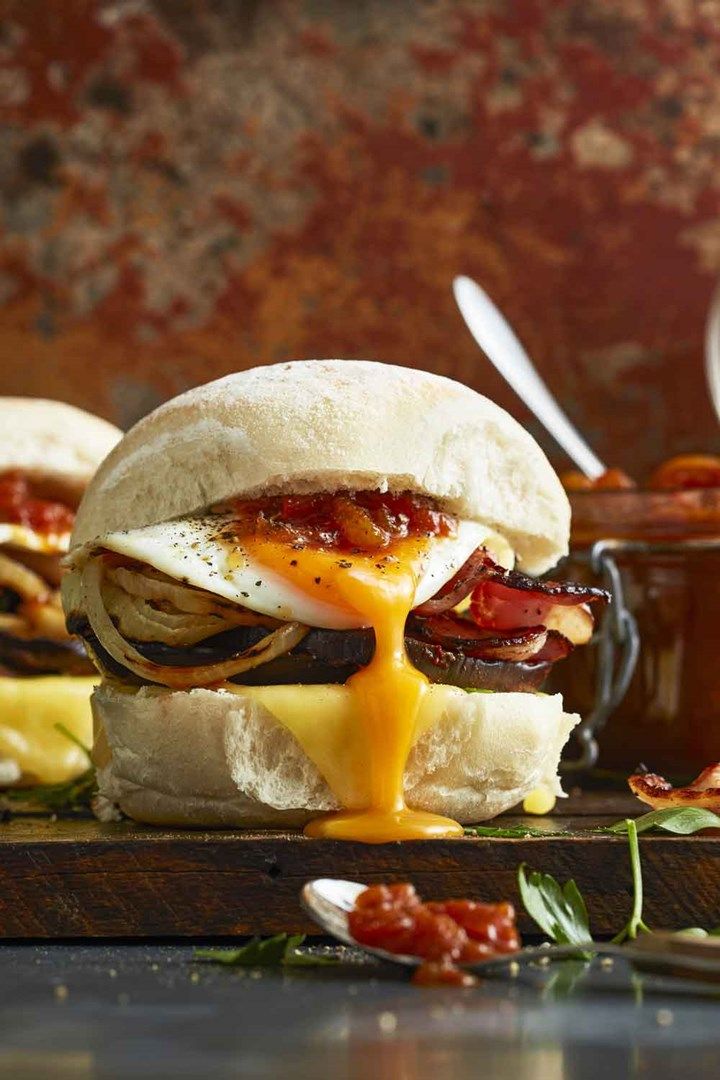 an egg sandwich with bacon and other toppings on a cutting board next to a bowl of sauce