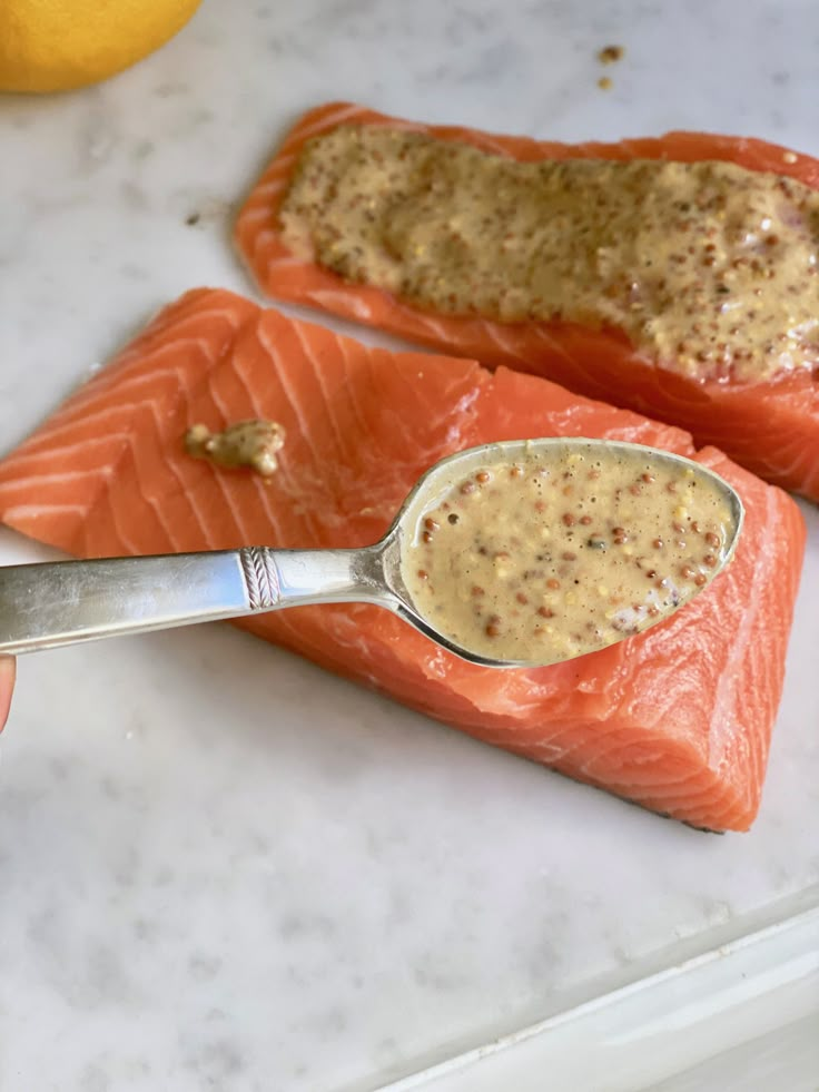 two salmon fillets with mustard being spooned into them on a marble counter top