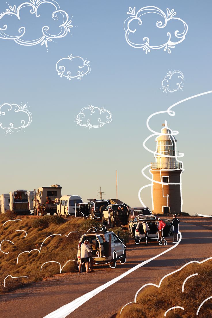 people standing on the side of a road next to cars and a light house with clouds in the sky