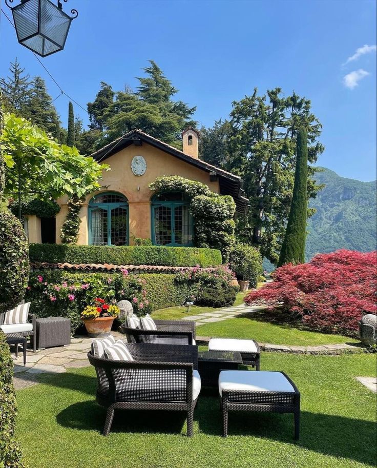 a patio with chairs and tables in front of a house surrounded by greenery, shrubs and flowers