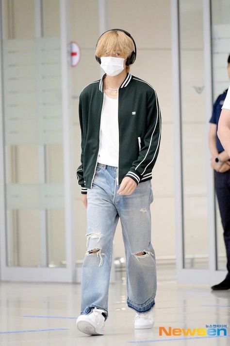 a man with headphones on walking in an airport wearing ripped jeans and a face mask