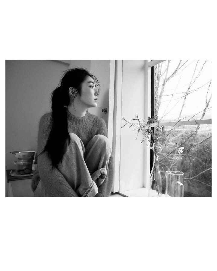 a black and white photo of a woman sitting on a window sill looking out the window