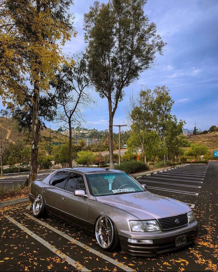 a silver car parked in a parking lot next to some trees and leaves on the ground