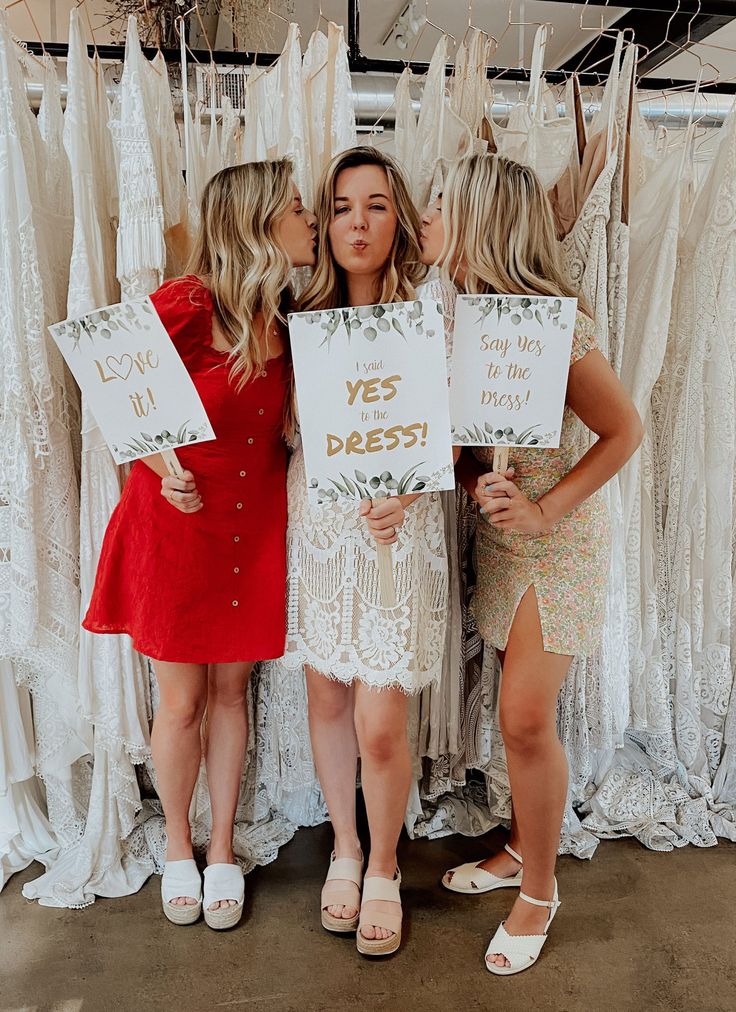 three women standing next to each other in front of white dresses and holding up signs