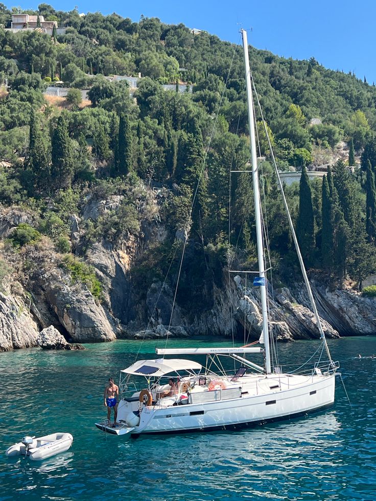 two people on a sailboat in the water near some hills and trees with houses