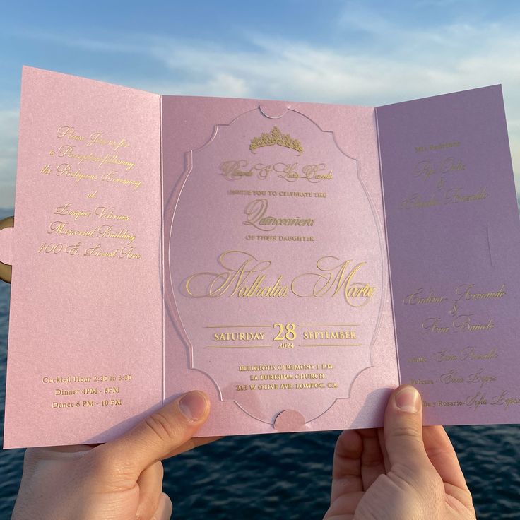 a person holding up a pink and gold wedding card in front of the ocean with blue sky