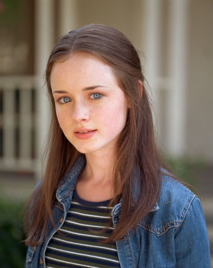 a young woman with long hair and blue eyes is looking at the camera while standing in front of a house