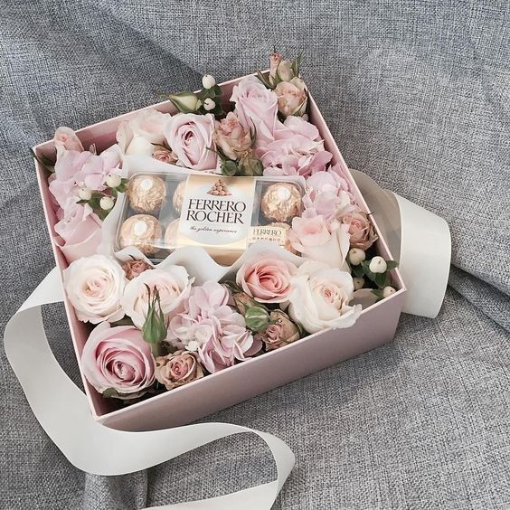 a pink box filled with roses and chocolates on top of a gray couch next to a white ribbon