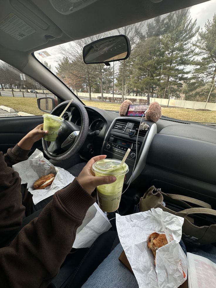 two people sitting in the drivers seat of a car eating food and drinking juice from cups