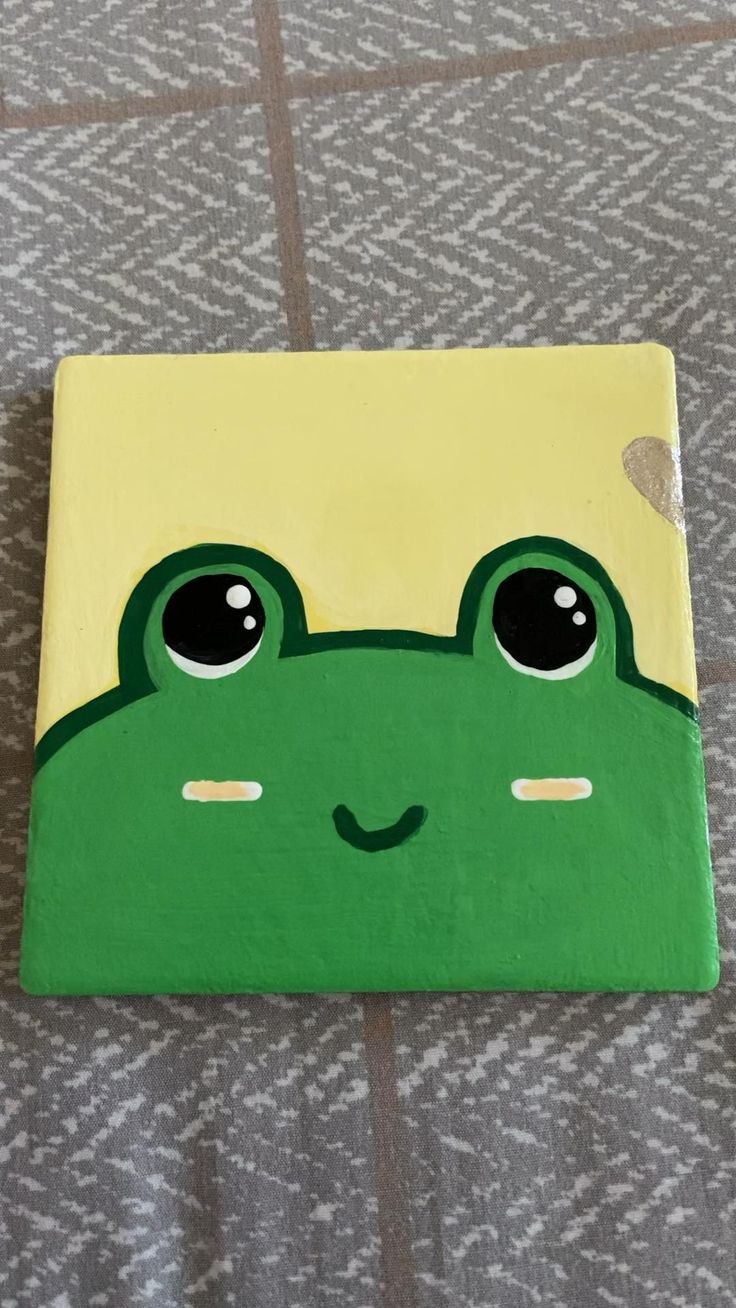 a green frog painted on top of a piece of wood with black eyes and nose