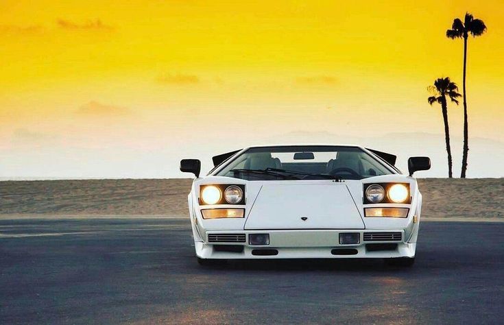 a white sports car is parked in the parking lot at sunset with palm trees behind it