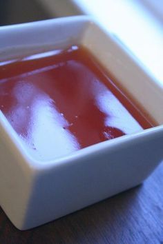 a white square bowl filled with red liquid on top of a wooden table next to a window