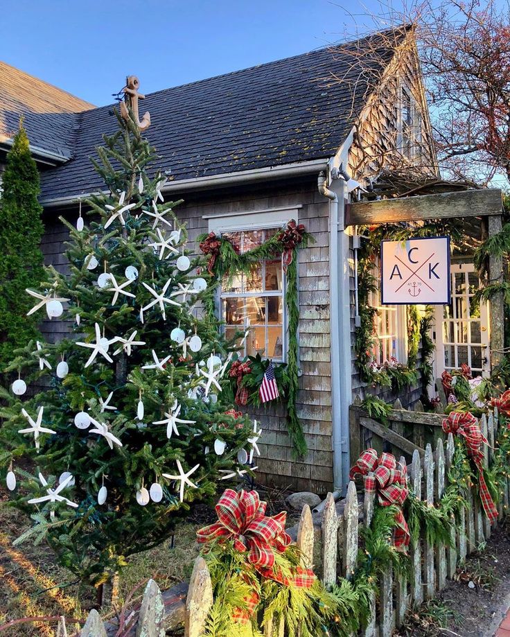 a christmas tree is in front of a small house with holiday decorations on the fence