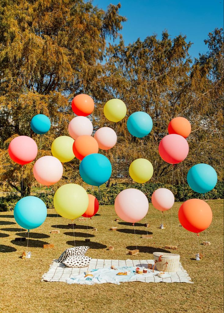 a bunch of balloons floating in the air on top of a grass covered field next to a picnic table