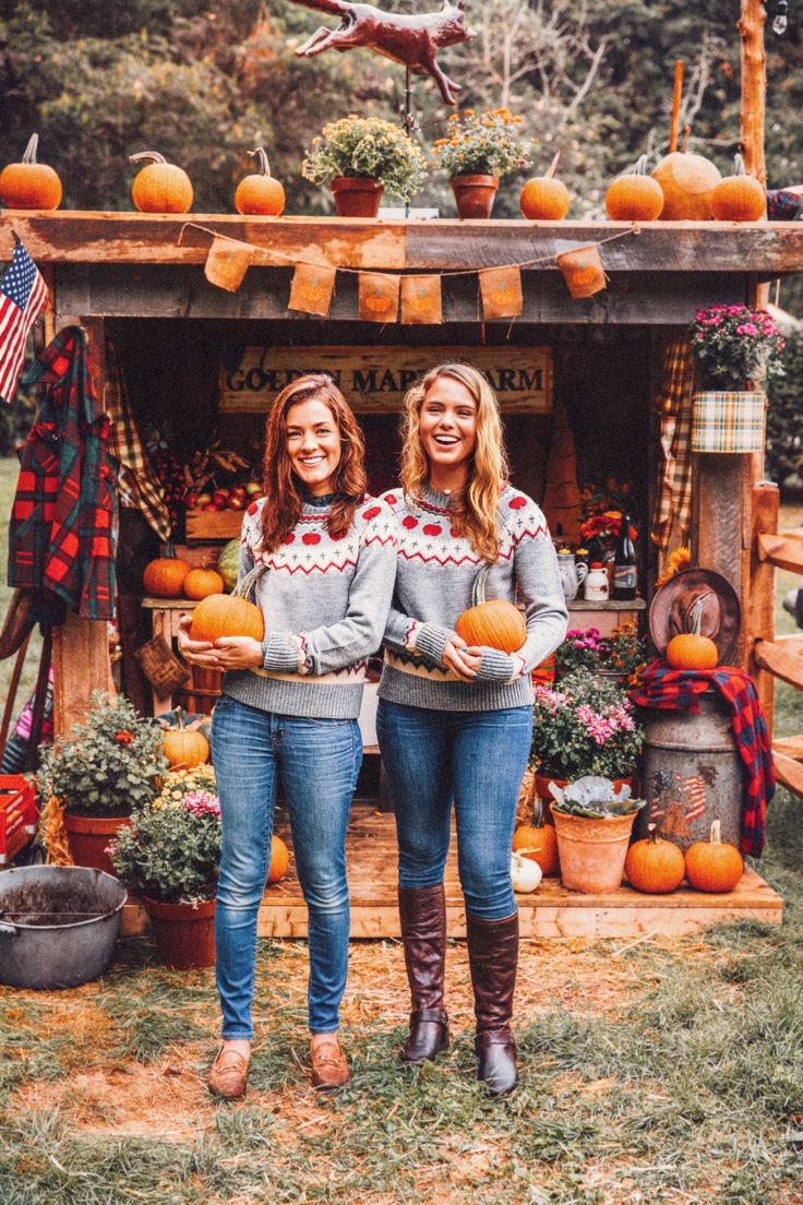 two women holding pumpkins in front of an outdoor stand with decorations on the roof