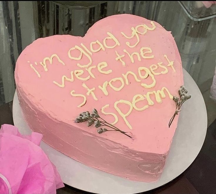 a pink heart shaped cake with writing on the side and flowers in front of it