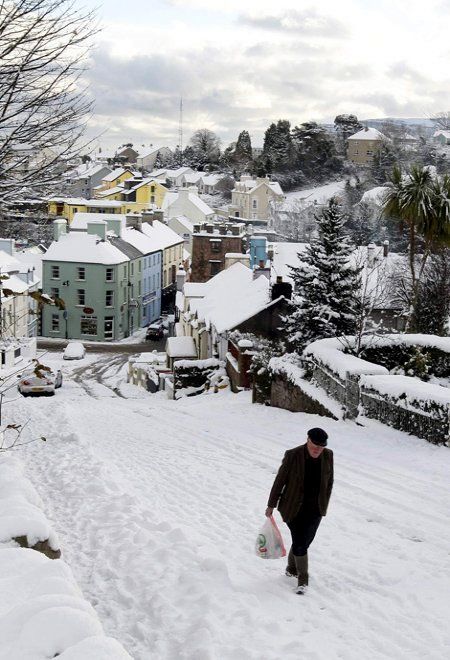 a man is walking through the snow with his skateboard in hand and buildings behind him