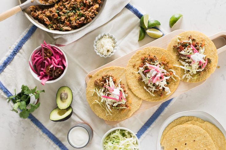 three tacos on a cutting board next to bowls of coleslaw and avocado