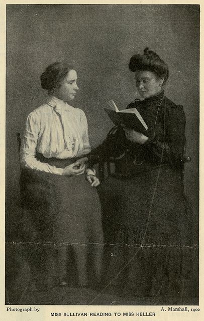 two women standing next to each other in front of a black and white photo, one reading a book