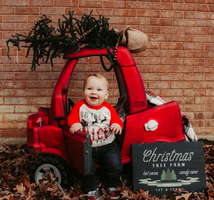 a baby sitting in a red truck with a christmas tree on the roof and sign next to it