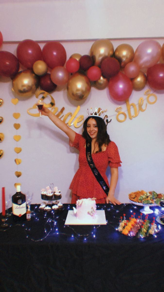 a woman standing in front of a table with cake and balloons on the wall behind her