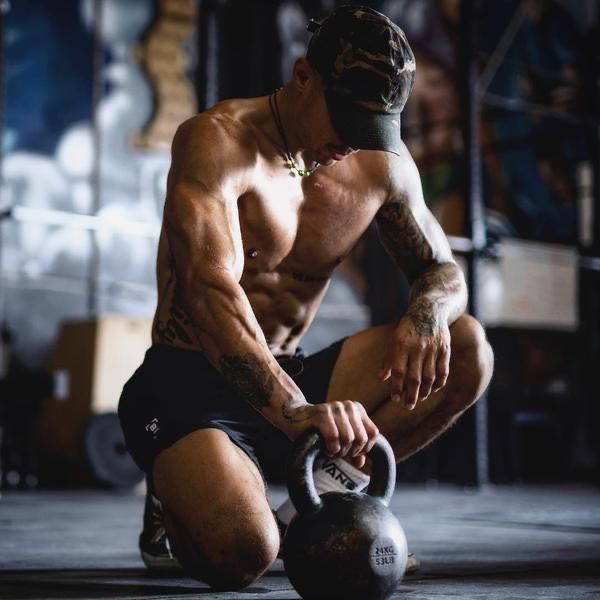 a shirtless man squatting down while holding a kettle in his right hand and looking at the ground