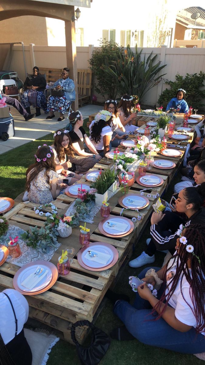 a group of people sitting around a wooden table with plates and flowers on it in the grass