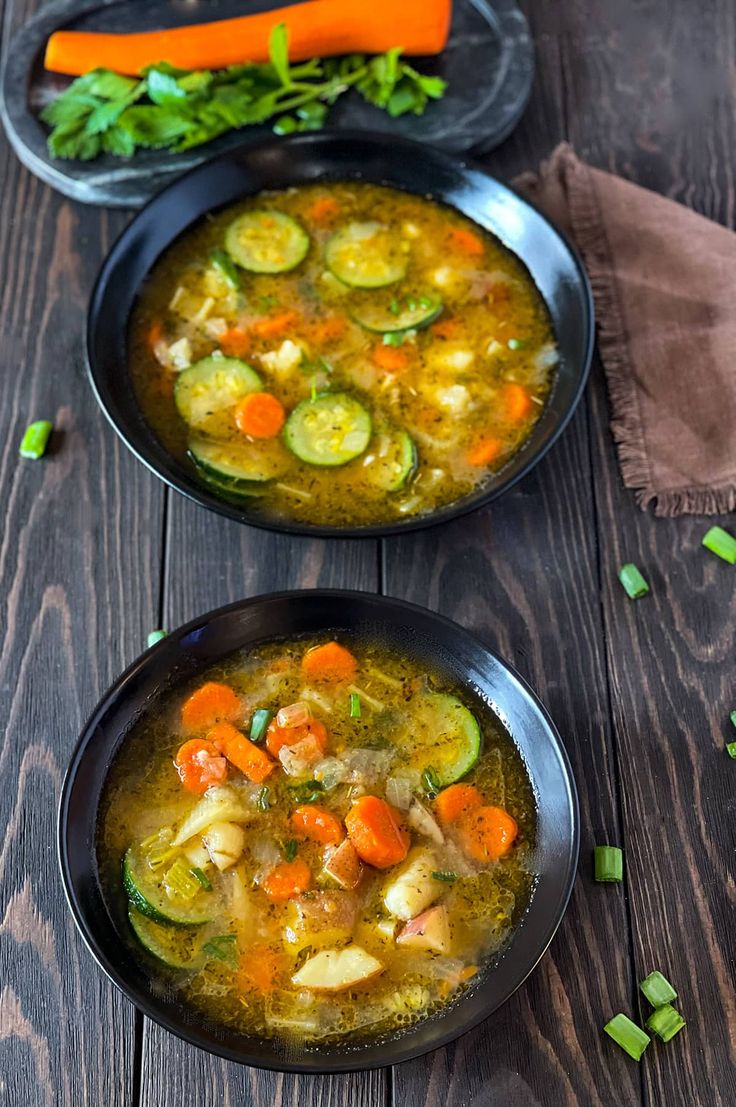 two black bowls filled with vegetable soup on top of a wooden table next to carrots and celery