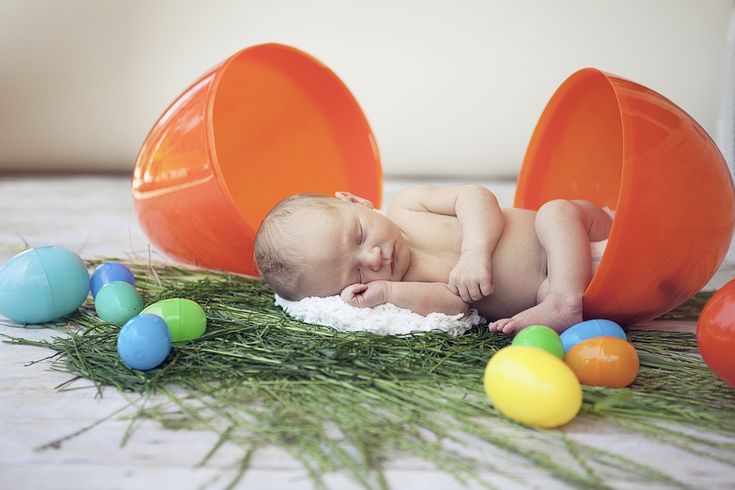 a baby laying on top of an orange bucket surrounded by easter eggs and fake grass