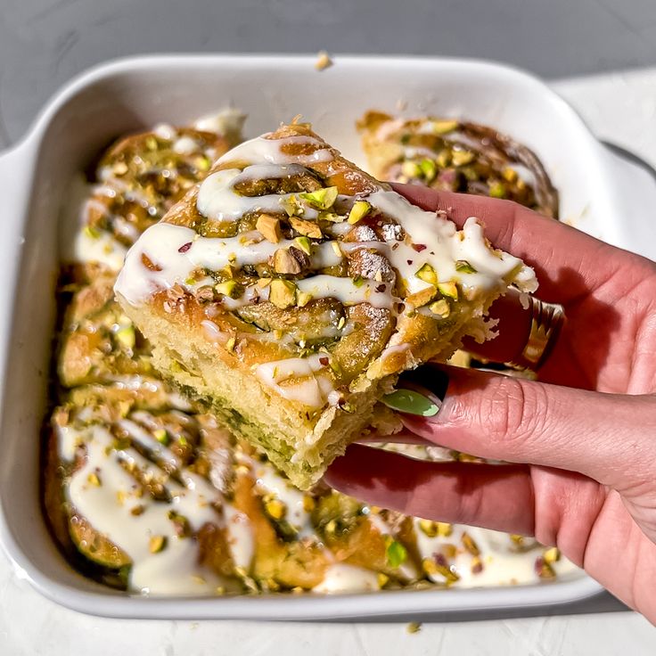 a hand holding up a piece of cake in a white casserole dish with icing and pistachio sprinkles