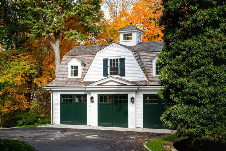 a white house with green garage doors in front of trees and leaves on the ground