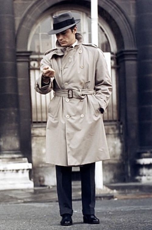 a man in a trench coat and hat standing on the street with his hands in his pockets