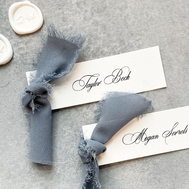 two place cards with ribbons tied to them on a gray table cloth next to buttons