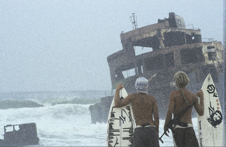 two surfers standing on the beach with their surfboards in front of an old ship