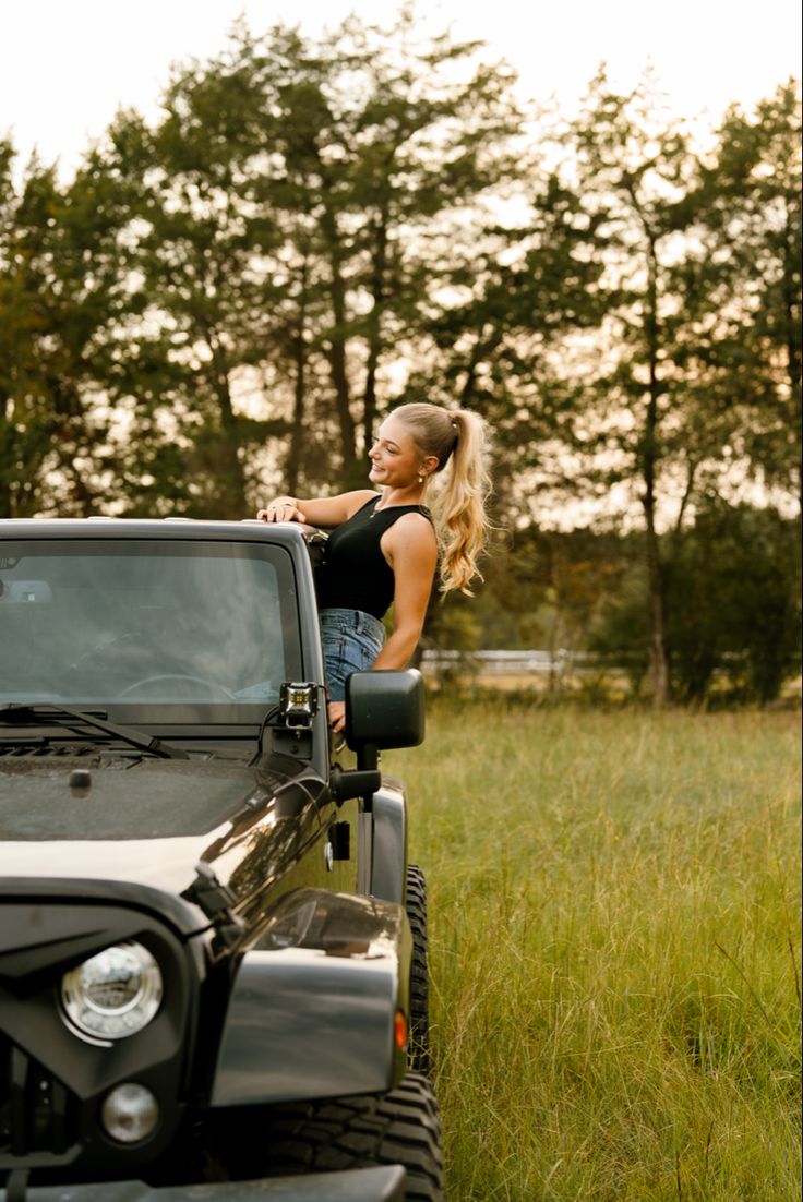 a woman leaning on the hood of a black jeep parked in a grassy field next to trees