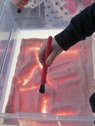 a child's hand holding a red marker over pink sand in a plastic container