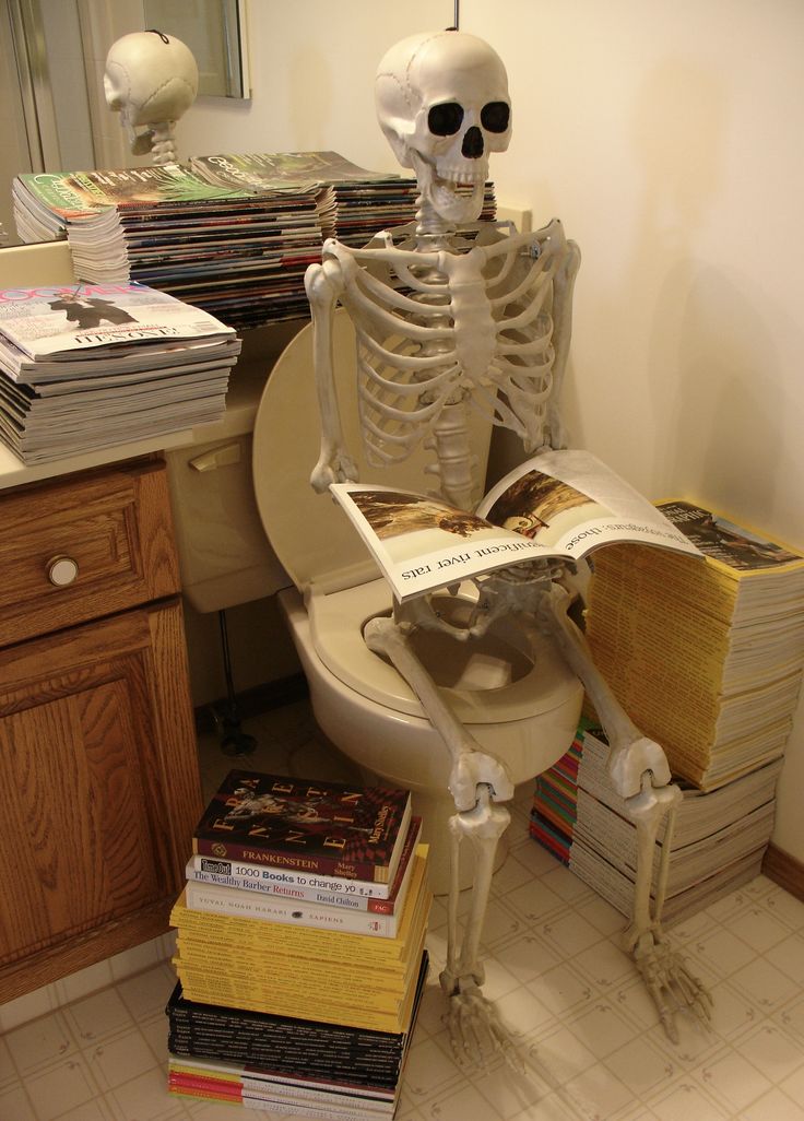 a skeleton sitting on top of a toilet reading a book next to stacks of books