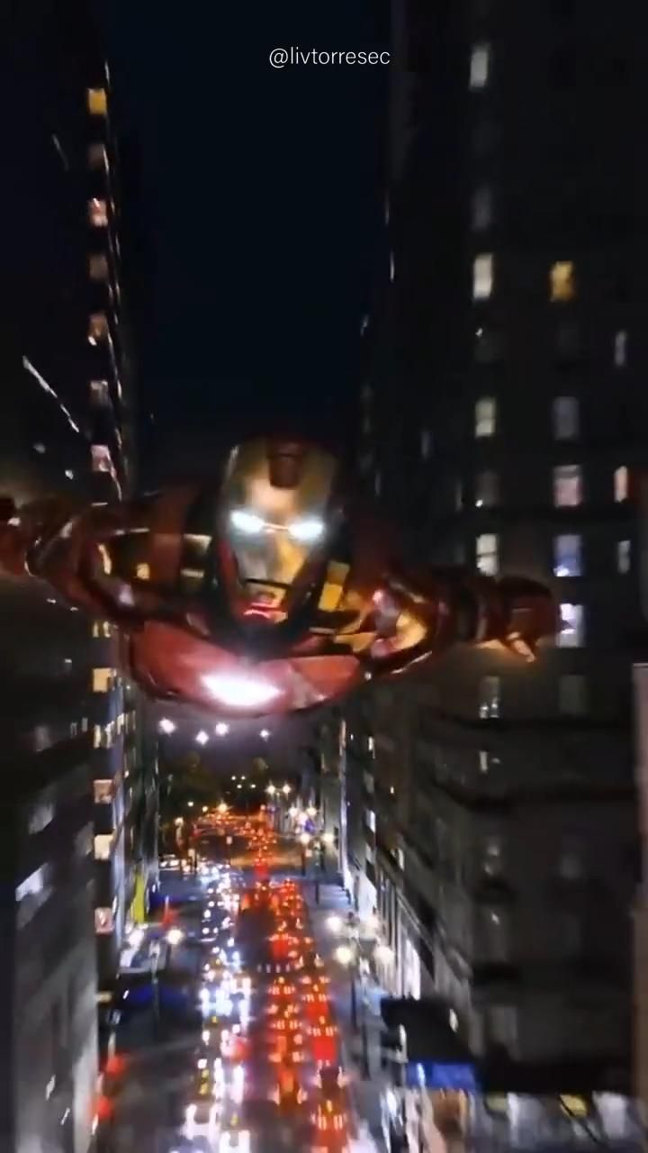 an iron man flying over a city at night with lots of traffic in the foreground