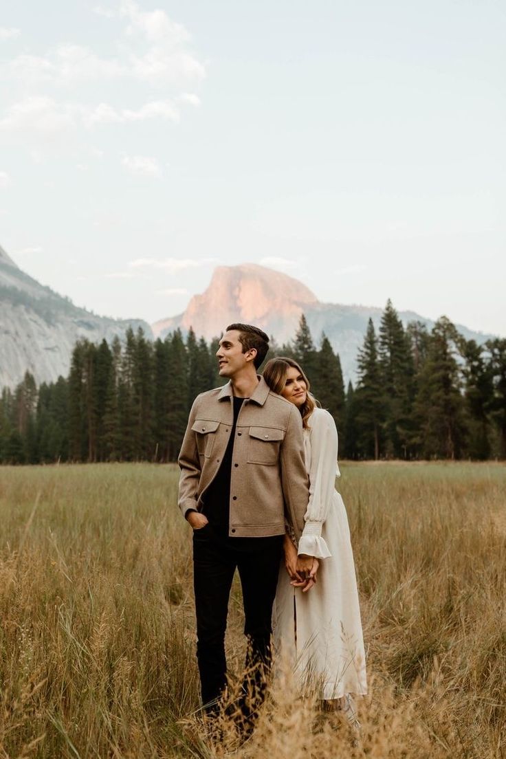 a man and woman are standing in tall grass with mountains in the backgroud