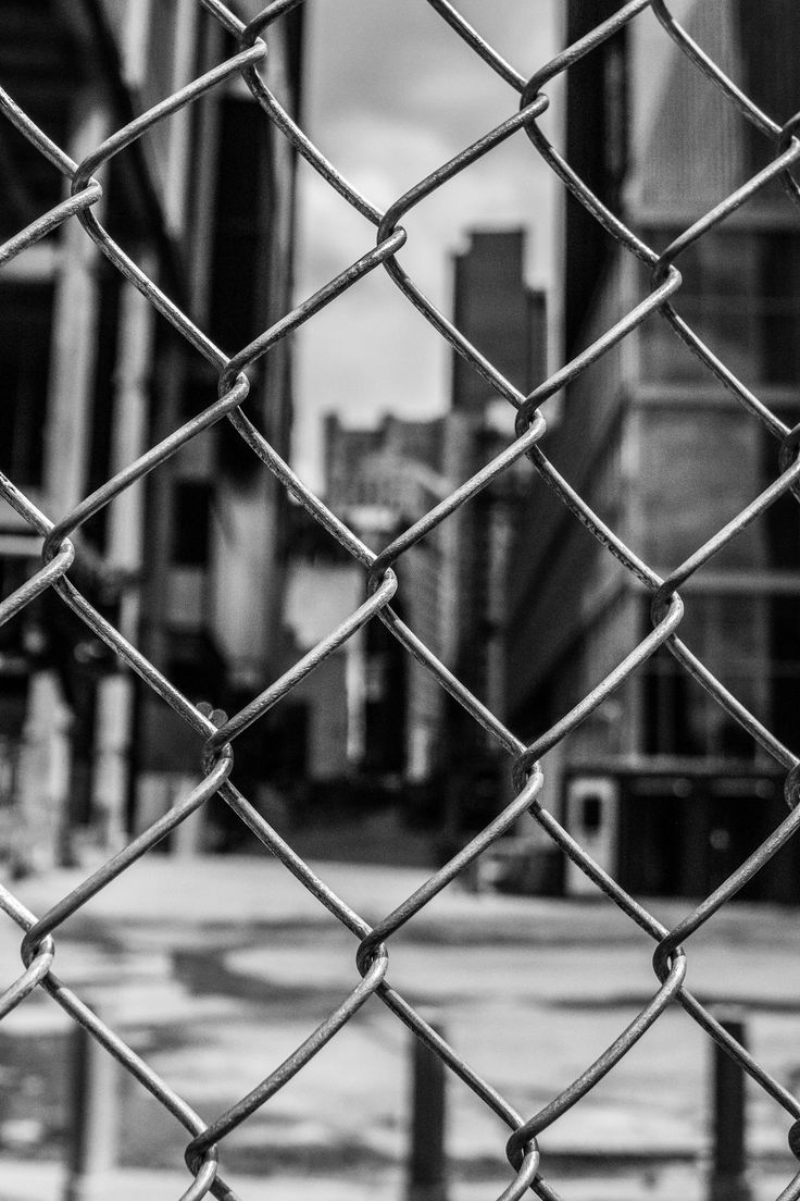 black and white photograph of a chain link fence with buildings in the distance behind it